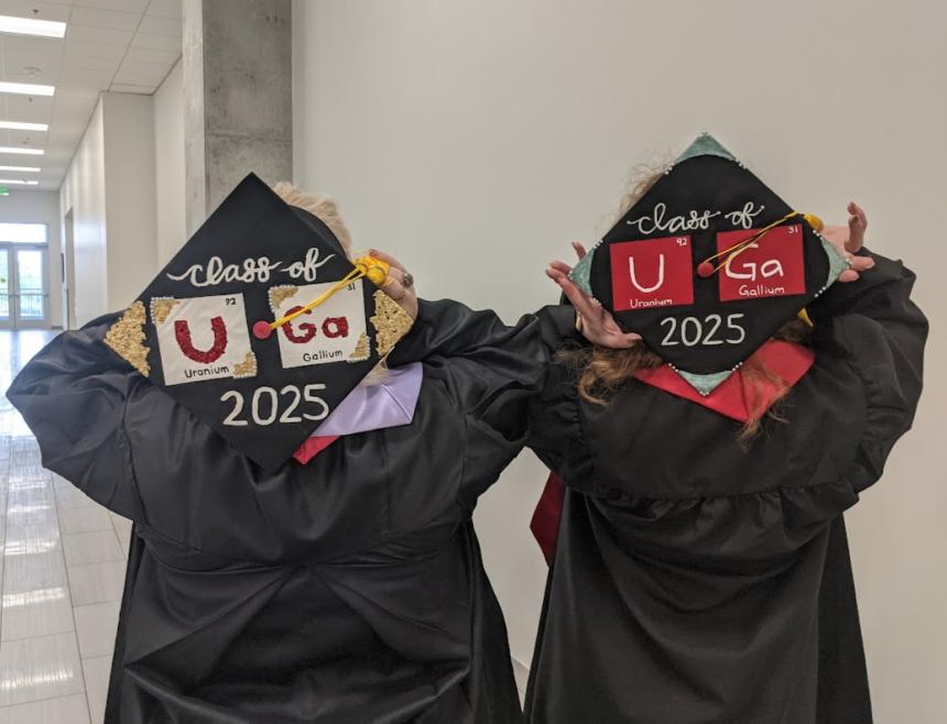 Image depicts two graduates in cap and gown with their backs to the camera, displaying decorated caps that read "UGA 2025," using the symbols for uranium and gallium to spell "UGA."