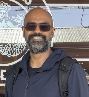 Portrait of Dr. Abishek K. Iyer, speaker, young man with dark hair and beard, standing in front of a coffee shop window