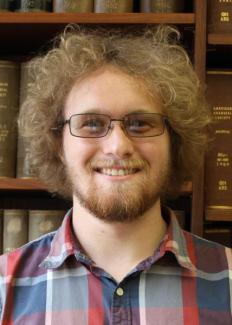 Headshot of Jason Colley, white male with curly blond hair, beard, and glasses; background of books in a library