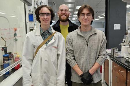 High school student Copper Callahan, science teacher Robert Pulliam, and UGA Chemistry graduate student Skyler Hollers stand in an organic chemistry lab with fume hood and lab bench in the background