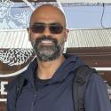 Portrait of Dr. Abishek K. Iyer, speaker, young man with dark hair and beard, standing in front of a coffee shop window