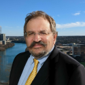 Head shot of bearded man with glasses; river and partial skyline in the background